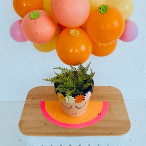 May include: A terracotta flower pot with a painted face and a flower crown, decorated with small white flowers. A green fern plant is growing in the pot. The pot is sitting on a wooden surface with a pink and orange half-circle underneath. A cluster of orange, pink, and yellow balloons with white and green flower accents are floating above the pot.