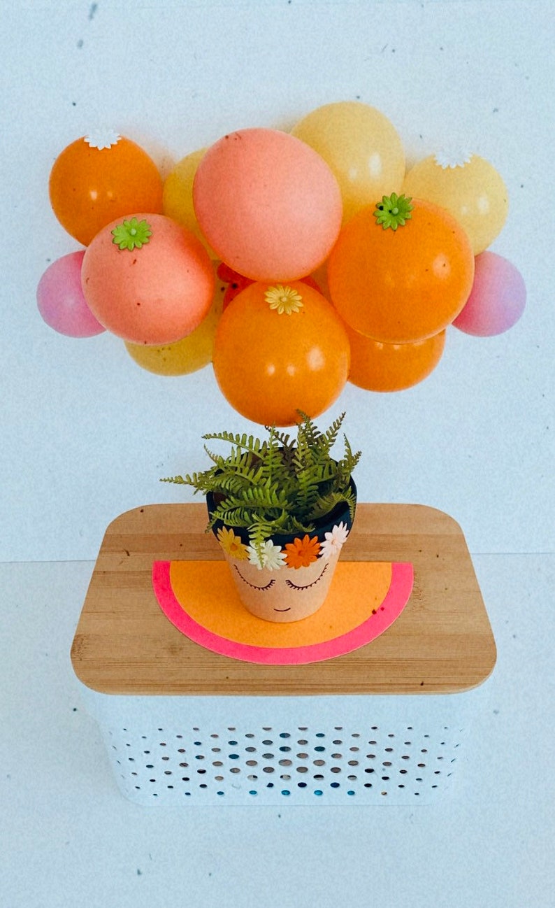 May include: A terracotta flower pot with a painted face and a flower crown, decorated with small white flowers. A green fern plant is growing in the pot. The pot is sitting on a wooden surface with a pink and orange half-circle underneath. A cluster of orange, pink, and yellow balloons with white and green flower accents are floating above the pot.