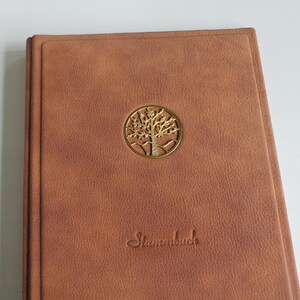 Könnte beinhalten: Ein braunes, ledergebundenes Buch mit einem goldenen Baumemblem in einem Kreis. Das Wort "Stammbuch" ist in kursiver Schrift unter dem Emblem geprägt. Das Buch hat eine strukturierte Oberfläche und eine rechteckige Form.