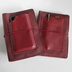 Könnte beinhalten: Zwei weinrote Lederjournale mit einer Tasche und einem elastischen Verschluss. Ein Journal enthält ein Smartphone, das andere einen Stift und eine herzförmige Büroklammer. Die Journale sind tiefrot.