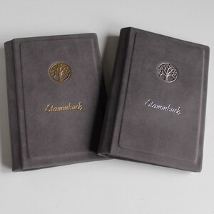 Könnte beinhalten: Zwei graue Bücher mit Soft-Touch-Oberfläche, jeweils mit einem kreisförmigen Baum-Emblem und dem Wort "Stammbuch" auf dem Einband. Ein Buch zeigt das Emblem und den Schriftzug in Gold, das andere in einer silbernen metallischen Ausführung.