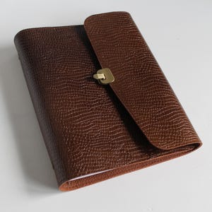 Könnte beinhalten: Ein braunes Lederjournal mit strukturierter Oberfläche und Klappenverschluss. Das Journal hat einen messingfarbenen Verschluss und einen abgerundeten Rücken. Das Cover hat eine satte, dunkelbraune Farbe.