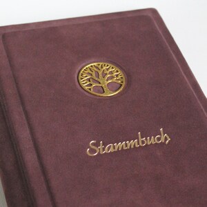 Könnte beinhalten: Ein dunkelviolettes Buchcover mit samtiger Textur und einem kreisförmigen, goldfarbenen Metallemblem, das einen Baum mit Ästen und Wurzeln zeigt. Das Wort "Stammbuch" ist in goldener Schrift unter dem Baumemblem auf dem Cover geprägt.
