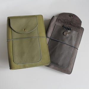 Könnte beinhalten: Zwei Leder-Notizbuchhüllen. Eine ist olivgrün mit einer Klappe und einem Knopfverschluss. Die andere ist grau mit einer Klappe und einem Knopfverschluss. Beide haben ein Gummiband zum Verschließen. Die Hüllen liegen auf einer weißen Oberfläche.