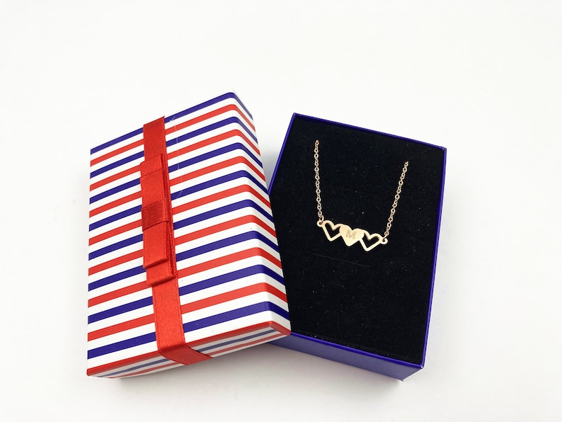 May include: A gold necklace with three connected heart charms in a blue and white striped gift box with a red ribbon.