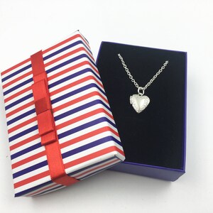 Puede incluir: Un collar con un dije en forma de coraz&oacute;n plateado en una caja de regalo a rayas rojas, blancas y azules con un lazo rojo.