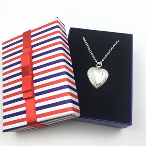 Puede incluir: Un collar con un dije en forma de coraz&oacute;n plateado en una caja de regalo a rayas azules y blancas con una cinta roja.