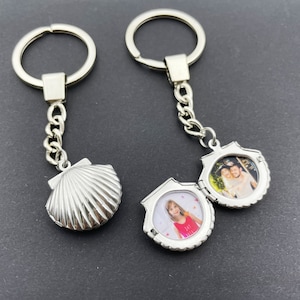 May include: Two silver keychains with photo locket charms. One charm is a seashell design, the other is a heart shape with a photo of a girl. Both charms are open to reveal a space for a photo.