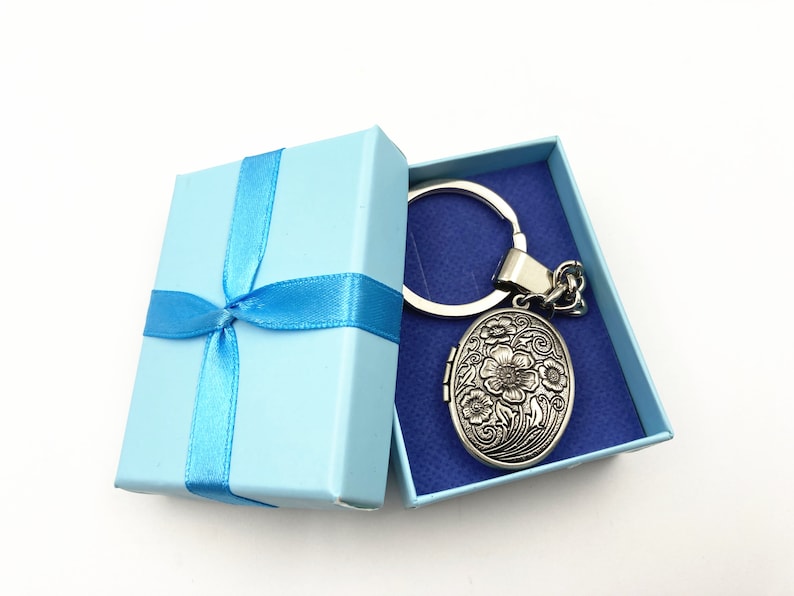 Puede incluir: Un llavero de plata con un medall&oacute;n con dise&ntilde;o floral se presenta en una caja de regalo azul claro con una cinta azul.