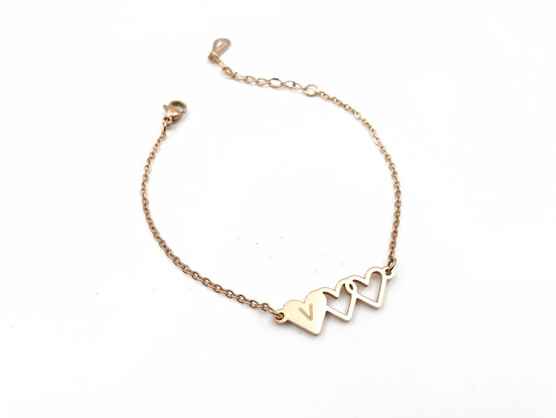 May include: Gold chain bracelet with three connected heart charms. The center heart charm has a small letter 'V' engraved inside.