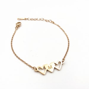 May include: Gold chain bracelet with three heart charms. The middle heart charm has the letter "M" engraved on it.