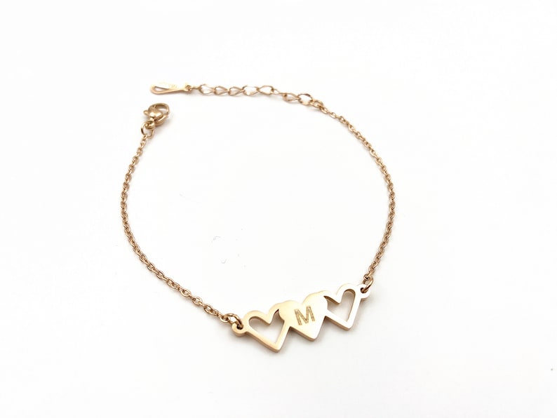 May include: Gold chain bracelet with three heart charms. The middle heart charm has the letter "M" engraved on it.