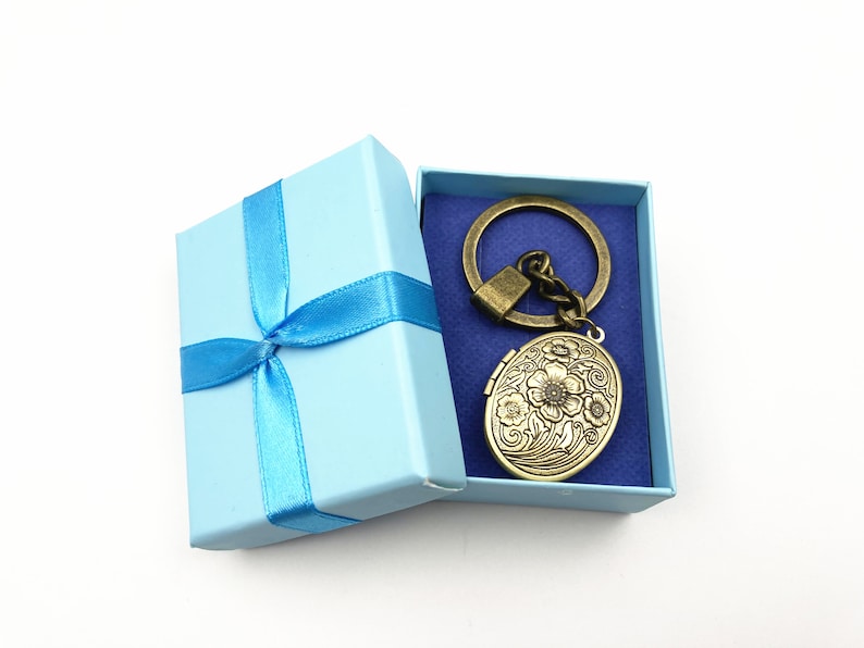 Puede incluir: Un medall&oacute;n ovalado dorado con dise&ntilde;os florales est&aacute; unido a un llavero. El medall&oacute;n est&aacute; en una caja de regalo azul claro con una cinta azul.