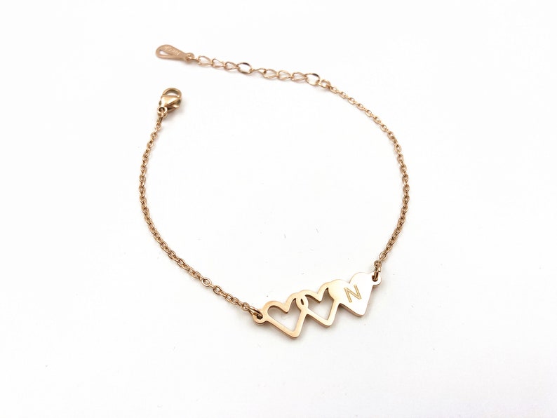 May include: A gold chain bracelet with three interlocking heart charms. The charm on the right has the letter "N" engraved on it.
