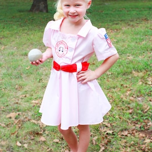 May include: A young girl wearing a pink baseball uniform with a red belt and a red bow in her hair. She is holding a white baseball and smiling. The uniform has the text "City of Rockford Peaches" on the front.