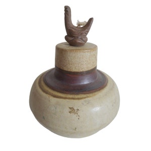 Puede incluir: Una lámpara de aceite de cerámica con una tapa en forma de pájaro. La lámpara está decorada con rayas marrones, rojas y blancas.