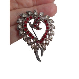 Puede incluir: Un broche de corazón de plata con diamantes de imitación rojos y transparentes. El broche tiene un diseño delicado y de hojas alrededor del corazón.