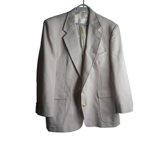 May include: A beige blazer hanging on a blue hanger. The blazer has two front pockets, gold buttons, and a notched lapel. The interior lining is a lighter shade of beige.