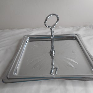Pode incluir: Uma bandeja de servir prateada com um design gravado decorativo e uma alça no centro. A bandeja é de forma quadrada e tem uma borda elevada.