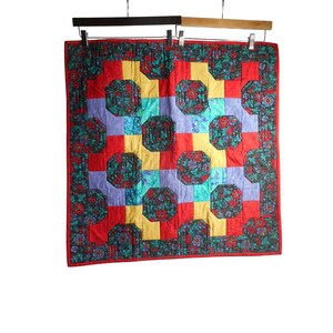 Op de afbeelding: Een rood, geel, blauw en groen patchwork-deken met een geometrisch patroon. Het deken is gemaakt van verschillende bloemenstoffen en heeft een zwarte rand.