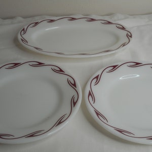Könnte beinhalten: Drei weiße Keramikteller mit einem burgunderfarbenen Blumendesign am Rand. Die Teller sind in verschiedenen Größen, darunter eine große ovale Platte und zwei kleinere runde Teller.