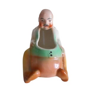 Könnte beinhalten: Vintage Keramikfigur eines sitzenden Mannes mit Glatze, Schnurrbart und rosigen Wangen. Die Figur trägt ein weißes Hemd, eine grüne Weste und eine orangefarbene Hose. Die Figur hat eine ausgehöhlte Mitte.