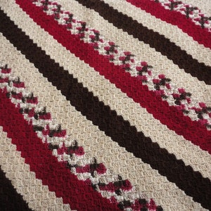 May include: A crocheted blanket with diagonal stripes in brown, beige, and red. The blanket has a textured pattern.