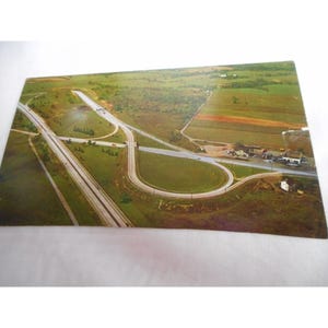 May include: An aerial view of a highway interchange with a cloverleaf design. The highway is surrounded by green fields and trees.