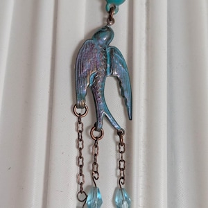 May include: A teal-colored bird pendant with a turquoise bead at the top and three dangling chains with blue crystal-like beads. The bird has iridescent blue and purple accents. The pendant is likely made of metal.