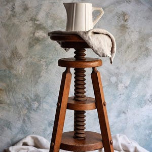 Puede incluir: Un taburete alto de madera con un mecanismo de tornillo central para ajustar la altura, con tres patas inclinadas y soportes circulares. Una jarra de cerámica color crema se asienta sobre el asiento, parcialmente cubierta por un paño de lino beige.