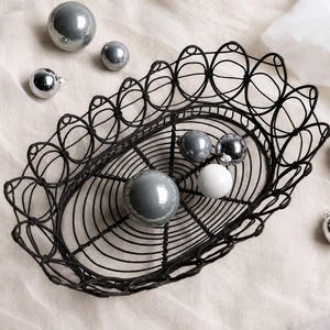 Può includere: Un cestino ovale in filo metallico nero con un bordo decorativo ad anelli, contenente diversi ornamenti rotondi e lisci in tonalità argento metallizzato, grigio e bianco. Altri ornamenti sferici abbinati sono sparsi attorno al cestino.