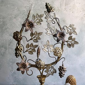May include: A brass-toned metal decorative stand featuring an ornate, wide base and a circular top frame adorned with sculpted metal flowers, leaves, and grape clusters. Two separate pinecone-shaped ornaments sit to the right of the stand.