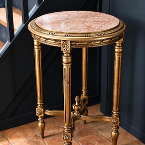 Op de afbeelding: Een ronde bijzettafel met een roze marmeren blad en een goudkleurig frame. De tafel heeft drie gecanneleerde poten die verbonden zijn door een centraal kruis met een decoratieve knop. Elke poot is versierd met uitgesneden bloemmotieven.