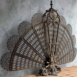 May include: A dark bronze-toned metal fireplace screen shaped like an open fan. The screen features a central vertical support, a decorative finial at the top, and a wide, semi-circular mesh panel with intricate, lace-like filigree edges.