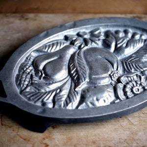 Może przedstawiać: Owalna, dekoracyjna taca z odlewanego metalu z szczegółowym reliefem gruszek, liści i kwiatów. Srebrna taca ma dwa małe uchwyty i spoczywa na drewnianej powierzchni. Projekt jest w stylu vintage i rustykalnym.