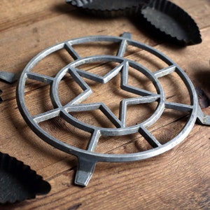 Może przedstawiać: Kolekcja zabytkowych narzędzi do pieczenia na drewnianej powierzchni. Centralnym elementem jest srebrny podkładka pod garnek z motywem gwiazdy. Wokół niego kilka małych, ciemnych metalowych foremek do tart, ukazujących rustykalną estetykę.