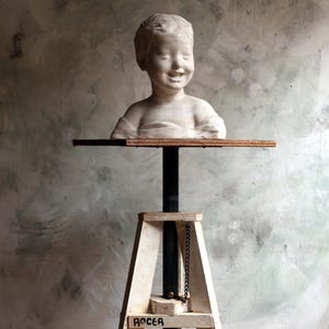 Op de afbeelding: Een wit gipsen borstbeeld van een kind met een lachende uitdrukking op een houten platform bovenop een hoge, witte, driepotige houten standaard. De standaard heeft een centrale zwarte metalen paal en een kleine metalen ketting, met de naam ROGER in zwart.