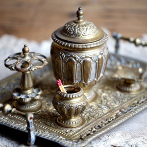 May include: A vintage brass smoking set with a tray, a match holder, a cigarette holder, and a lidded jar. The set is decorated with intricate floral patterns and is sitting on a lace doily.