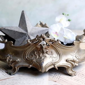 Op de afbeelding: Een sierlijke, antieke, goudkleurige metalen centerpiece kom met bloemen- en krulwerkdetails. Een grijze ster en witte orchideeën liggen in de kom. De kom heeft handvatten en staat op oude papieren.