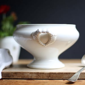 Può includere: Una zuppiera in ceramica bianca con manici decorativi, una ciotola larga e una base a piedistallo. Un mestolo d'argento poggia su una superficie di legno accanto alla zuppiera. Un panno bianco e un vaso con un fiore rosso sono sullo sfondo.