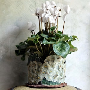 Puede incluir: Una planta de ciclamen blanca con hojas verdes en una maceta de cerámica cilíndrica con un relieve floral texturizado y pintado a mano en tonos verde, crema y marrón, sobre un pequeño taburete redondo de madera oscura con patas.