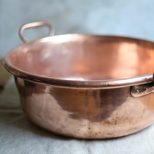 Puede incluir: Un cuenco de cobre con dos asas curvas. El cuenco tiene un diseño ancho y poco profundo y una superficie brillante y reflectante. El cobre tiene un color marrón rojizo cálido, y el cuenco parece ser vintage o antiguo.