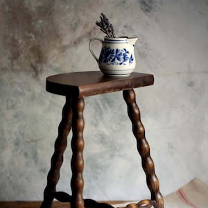 May include: A dark wooden stool with a round top and three twisted legs. A small, white jug with blue floral patterns holds dried lavender. The stool is a classic, rustic design.