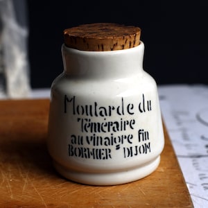 May include: A white ceramic mustard pot with a cork stopper. The pot has black text that reads "Moutarde du Téméraire au vinaigre fin BORMIER DIJON". The pot is on a wooden chopping board.