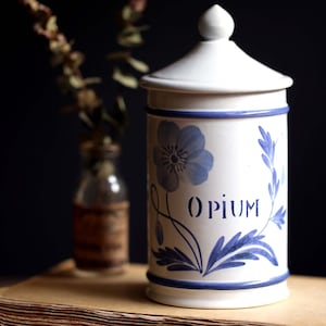 May include: A white ceramic apothecary jar with a lid, decorated with blue floral patterns and the word "OPIUM." The jar is set against a dark backdrop, suggesting a vintage style. A small glass bottle is in the background.