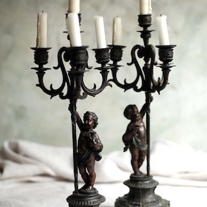 Puede incluir: Dos candelabros antiguos de bronce. Cada candelabro presenta una figura de querubín que sostiene un poste central, que soporta múltiples candeleros. Los candelabros tienen brazos ornamentados y enrollados y están rematados con velas blancas. La base está detallada con pies decorativos.