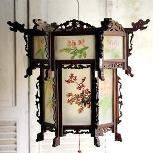 Może przedstawiać: Ozdobna, ciemna drewniana latarnia z malowanymi panelami kwiatowymi. Latarnia ma sześciokątny kształt z dekoracyjnymi rzeźbami i czerwonym frędzlem. Panele przedstawiają delikatne malowidła kwiatów i gałęzi.