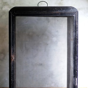 Puede incluir: Pantalla rectangular con marco negro y un interior de malla metálica. El marco tiene una parte superior redondeada con un pequeño asa. La pantalla está sobre una superficie de madera.