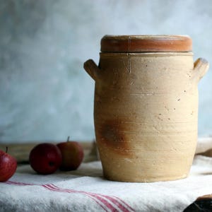 以下が含まれることがあります： 蓋付きのクリーム色のヴィンテージセラミックジャー。ジャーは素朴な手作りの外観で、シンプルな水平線の模様で飾られています。ジャーは赤色のストライプのある白いリネン製のテーブルクロスの上に置かれています。