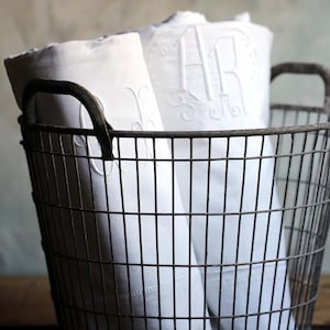 Puede incluir: Una cesta de alambre llena de ropa de cama blanca enrollada. La ropa de cama tiene iniciales bordadas. La cesta tiene dos asas y está sobre una superficie de madera. El fondo es gris apagado.